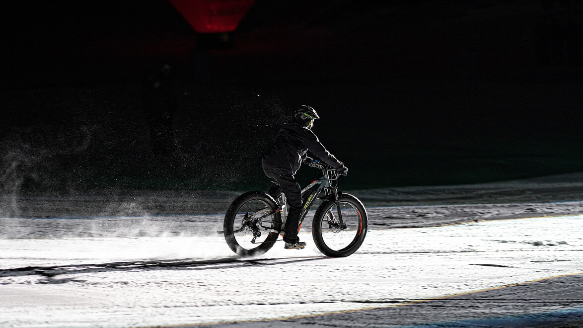 Eine Person fährt mit einem Fatbike auf der verschneiten Piste bei Dunkelheit. / A person rides a fatbike on a snowy slope in the dark.