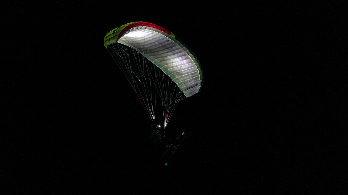 Ein Gleitschirmflieger mit leuchtendem Schirm ist bei Nacht über der Skipiste zu sehen. / A paraglider with an illuminated canopy is seen above the ski slope at night.