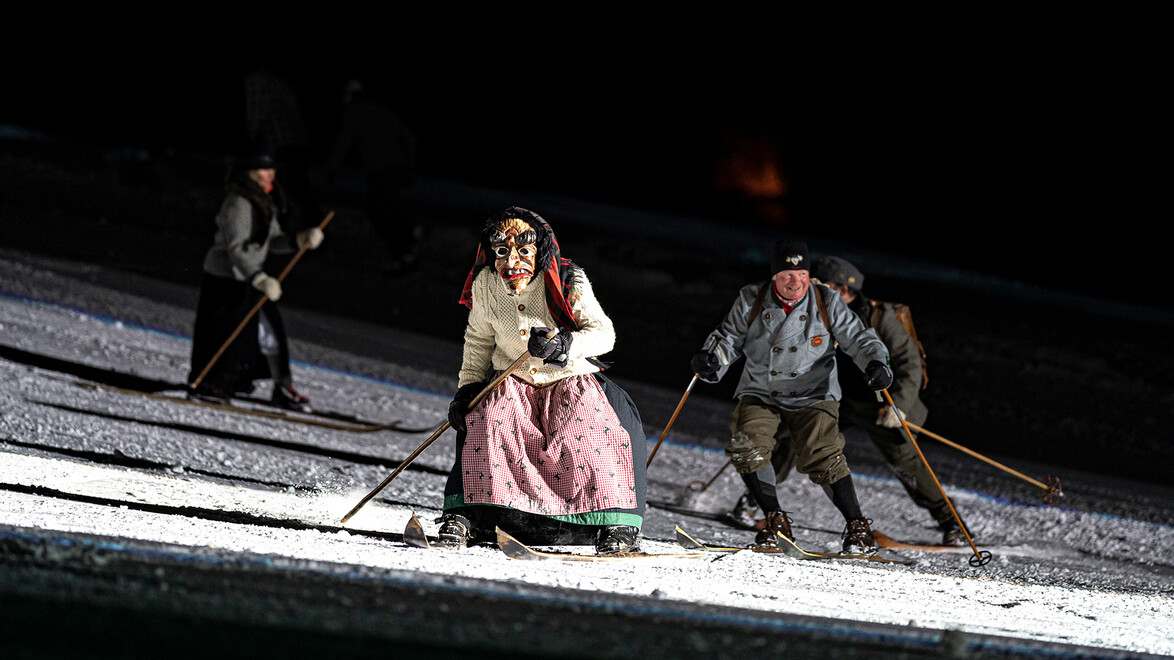 Historisch gekleidete Skifahrer:innen fahren bei Nacht über eine beleuchtete Piste. / Skiers in historical costumes descend a floodlit slope at night.