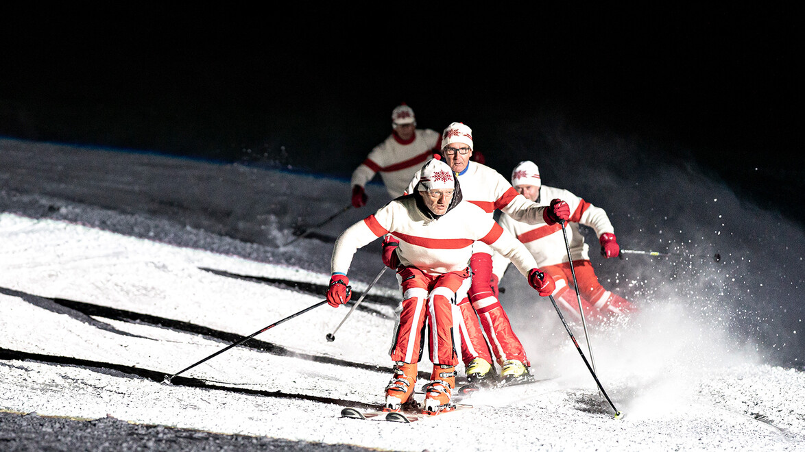 Fünf Skifahrer:innen in roten und weißen Outfits fahren synchron über die nächtliche Skipiste. / Five skiers in red and white outfits ski synchronously down the night slope.