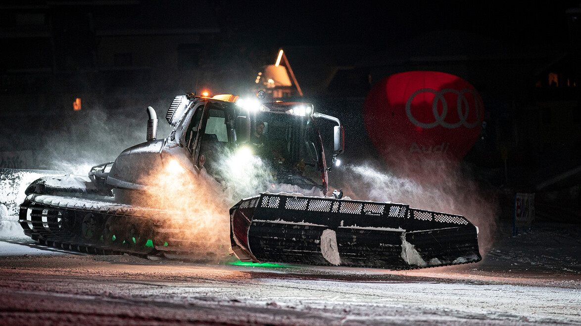 Ein Pistenfahrzeug präpariert nachts unter Flutlicht eine Skipiste, Schnee wirbelt auf. / A snow groomer prepares a ski slope at night under floodlights, snow spraying around.