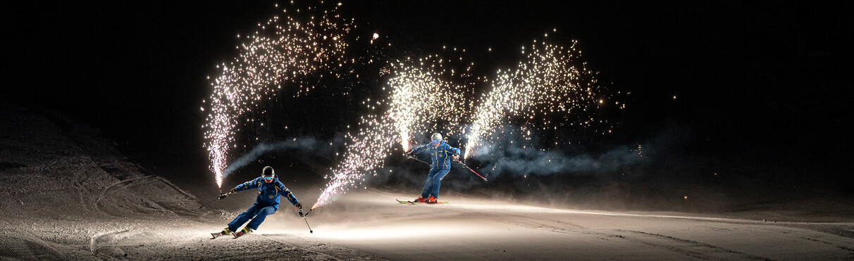 Zwei Skifahrer mit Funken-Fackeln gleiten bei Nacht über eine Piste.