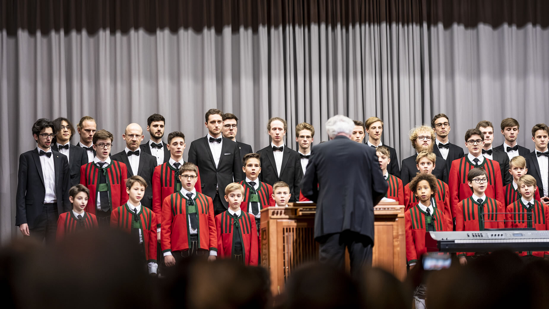 Ein Knabenchor und Männerchor singen gemeinsam unter der Leitung eines Dirigenten auf einer Bühne / A boys’ and men’s choir perform together under a conductor on stage