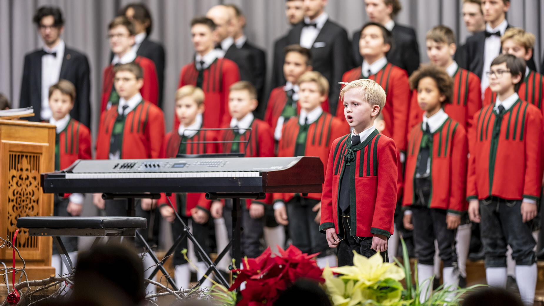 Ein Kinderchor in roten Uniformen singt auf der Bühne, im Vordergrund Blumen und ein Keyboard / A children’s choir in red uniforms sings on stage, with flowers and a keyboard in the foreground
