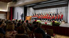 Publikum sitzt in einem Konzertsaal, während ein Knabenchor und ein Dirigent auf der Bühne musizieren / Audience seated in a concert hall, while a boys’ choir and conductor perform on stage