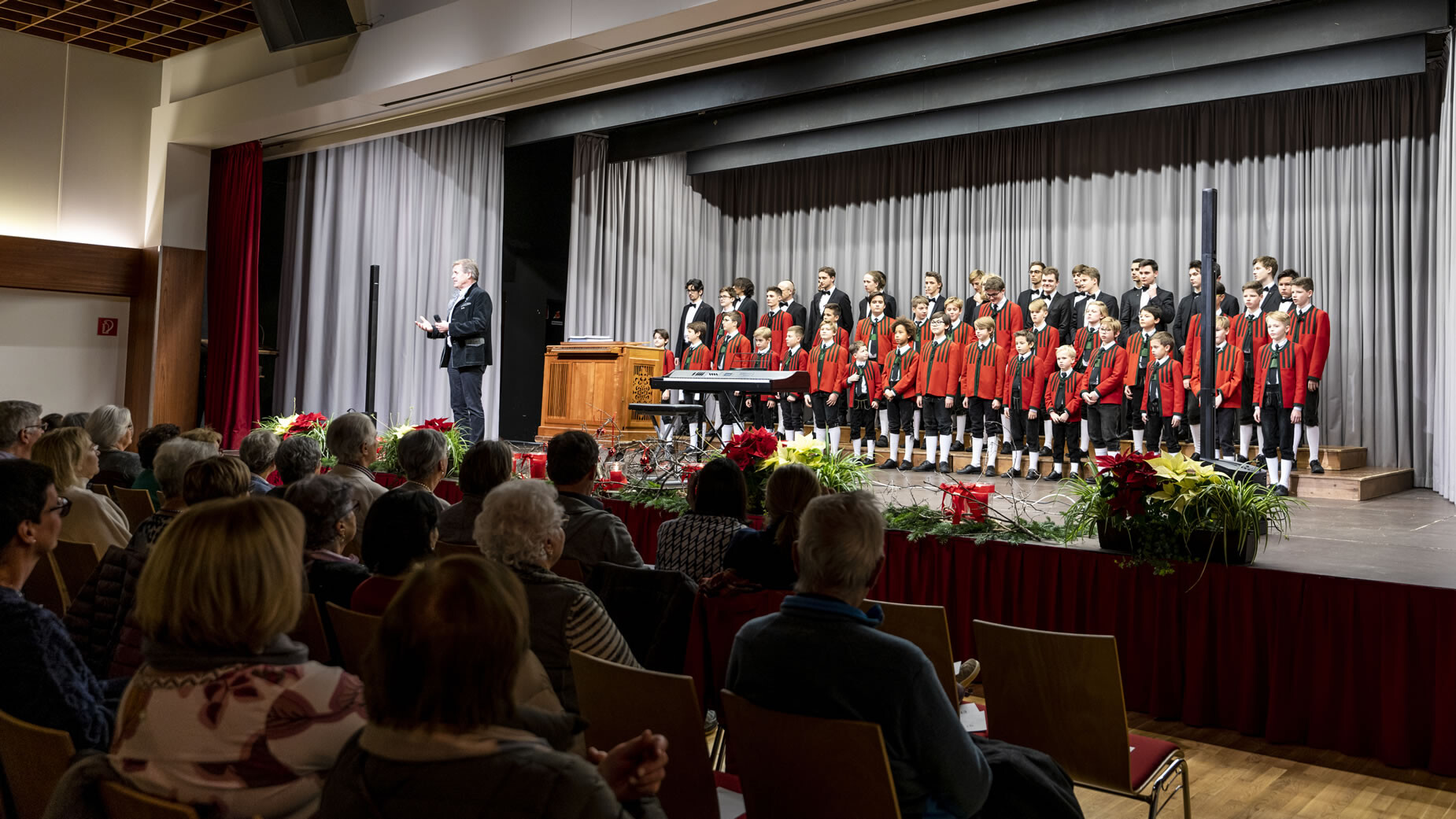 Publikum sitzt in einem Konzertsaal, während ein Knabenchor und ein Dirigent auf der Bühne musizieren / Audience seated in a concert hall, while a boys’ choir and conductor perform on stage