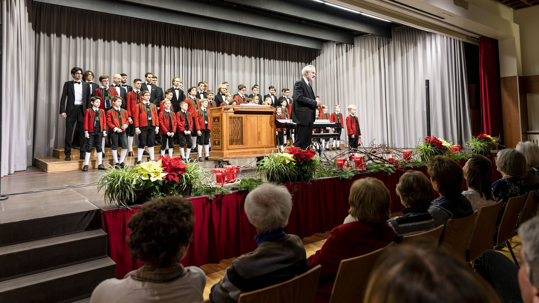Blick auf das Publikum und den Chor auf der Bühne mit Weihnachtsdekorationen im Vordergrund / View of the audience and choir on stage with Christmas decorations in the foreground