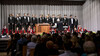 Ein Männerchor in schwarzen Smokings steht auf der Bühne, begleitet von einem Dirigenten am Pult / A men’s choir in black tuxedos stands on stage, led by a conductor at the podium