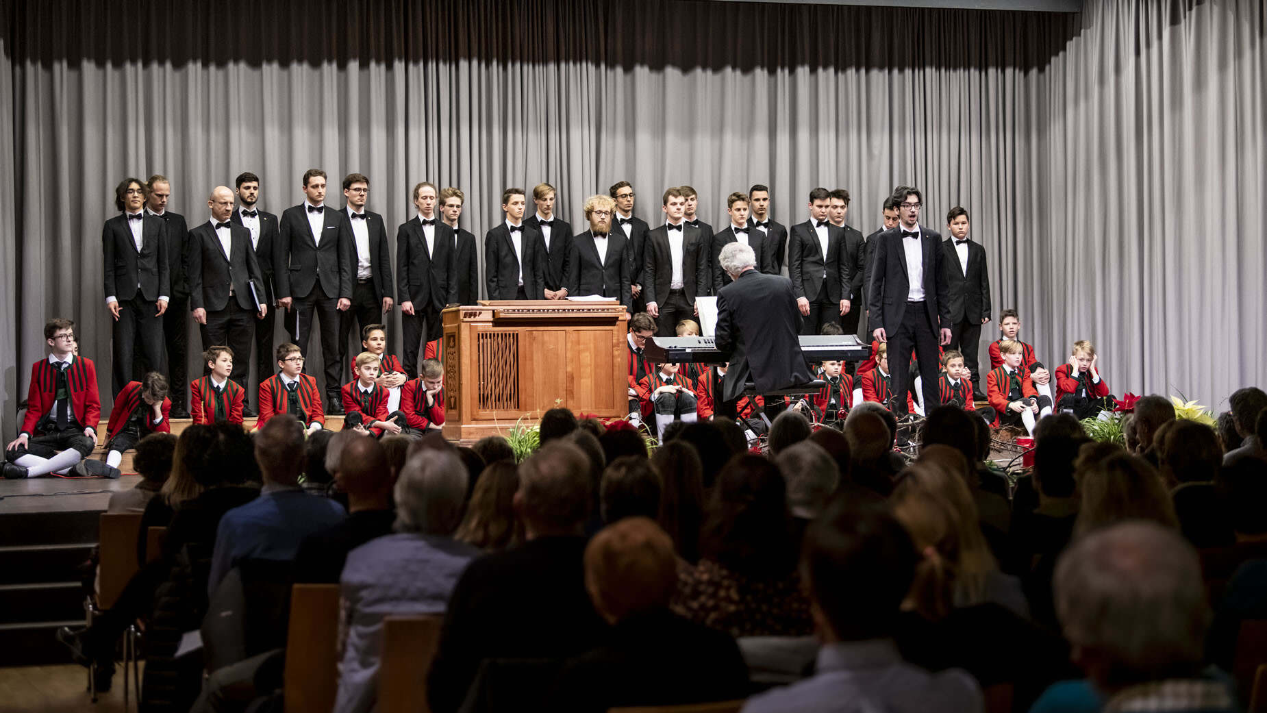 Ein Männerchor in schwarzen Smokings steht auf der Bühne, begleitet von einem Dirigenten am Pult / A men’s choir in black tuxedos stands on stage, led by a conductor at the podium