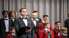 Ein Solist im Smoking steht vor einem Kinderchor und hält ein Notenblatt / A soloist in a tuxedo stands in front of a children’s choir holding sheet music
