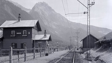 Historisches Foto des Bahnhofs Schnann mit Schienen und Bergen im Hintergrund.