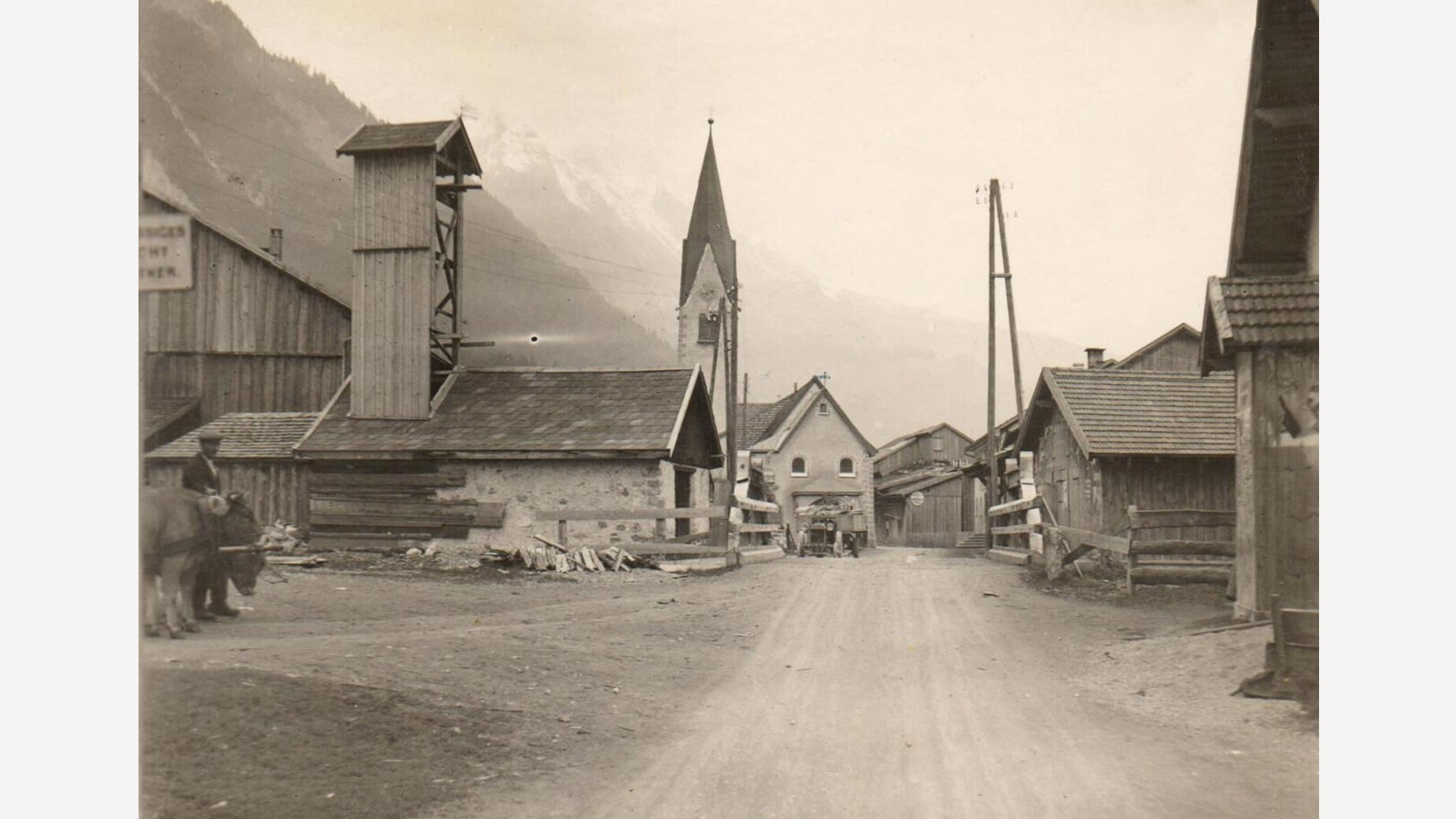 Alte Aufnahme mit Blick auf das Feuerwehrhaus und die Kirche von Schnann, flankiert von traditionellen Holzhäusern / Old photograph showing Schnann’s fire station and church, surrounded by traditional wooden houses