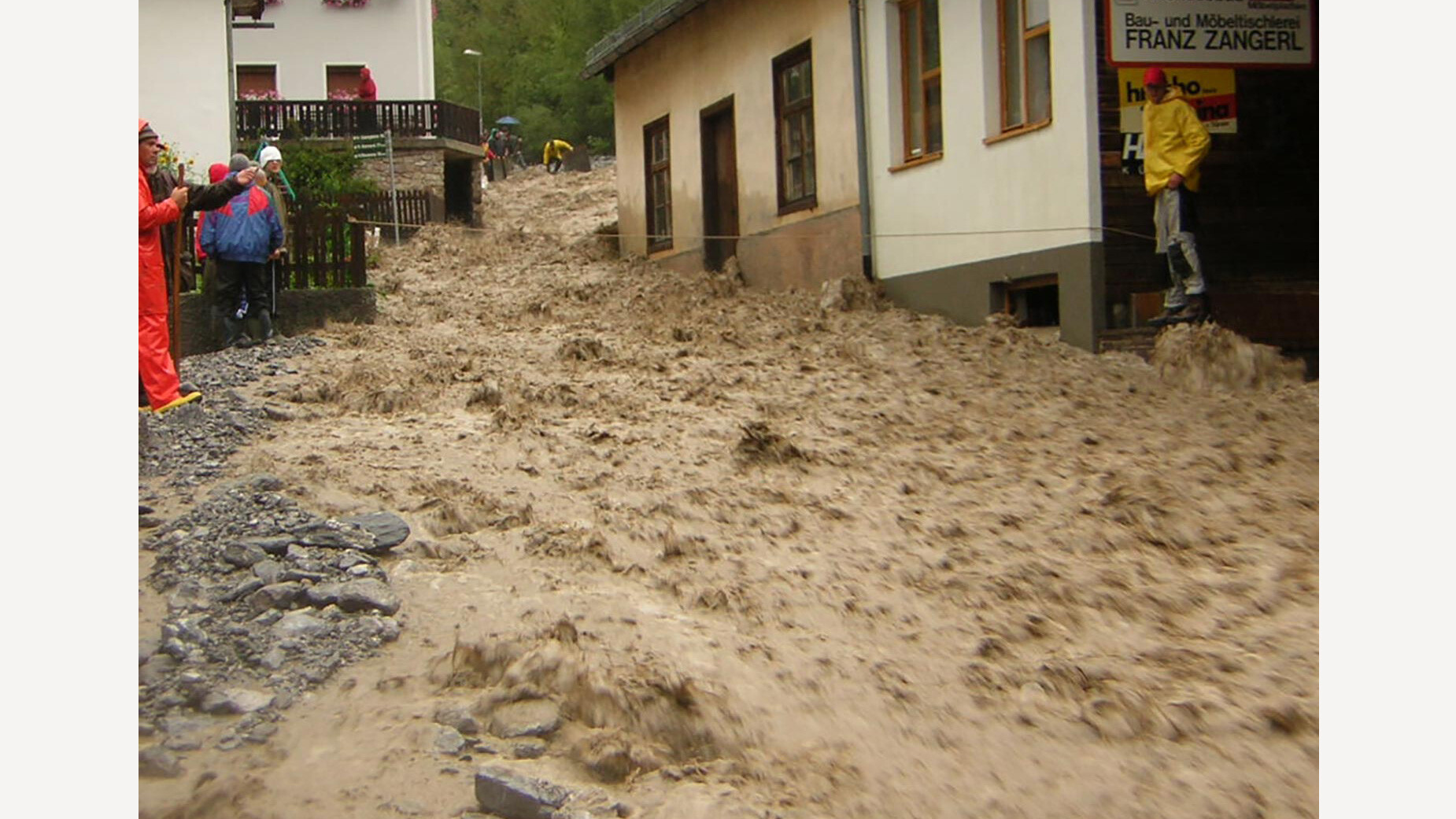 Heftige Mure wälzt sich durch Straße, Häuser links und rechts gefährdet / Intense mudslide surges through street, houses on both sides endangered