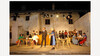 Theaterszene mit traditioneller Kleidung vor historischer Hausfassade, mehrere Darsteller:innen stehen und sitzen auf der Freilichtbühne / Theatre scene in traditional clothing in front of a historic house facade, several performers standing and sitting on the open-air stage