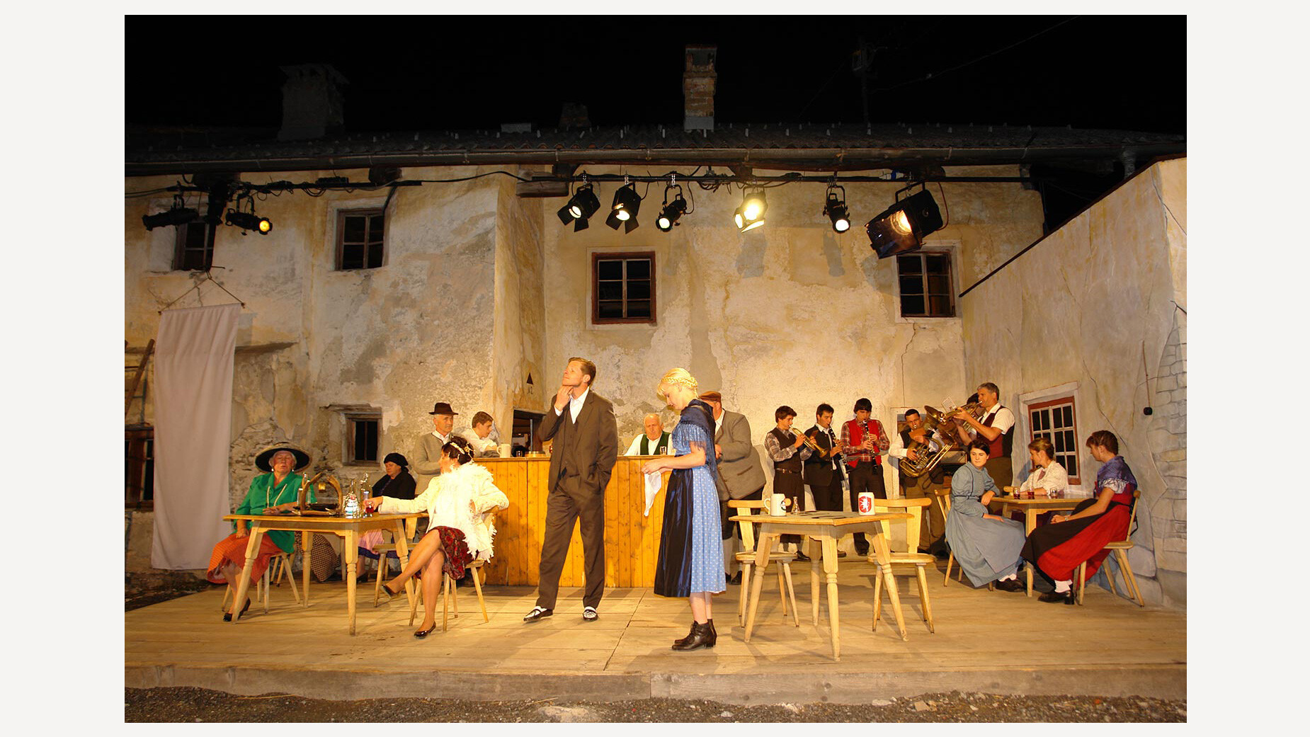 Theaterszene mit traditioneller Kleidung vor historischer Hausfassade, mehrere Darsteller:innen stehen und sitzen auf der Freilichtbühne / Theatre scene in traditional clothing in front of a historic house facade, several performers standing and sitting on the open-air stage