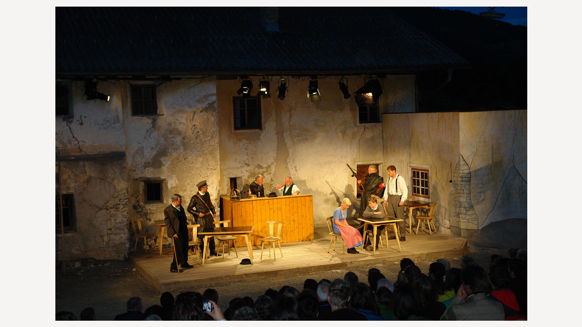 Abendliche Theateraufführung im Freien mit beleuchteter Kulisse und Publikum, Hauptdarsteller:innen agieren in der Mitte der Bühne / Evening outdoor theatre performance with illuminated backdrop and audience, lead actors performing center stage