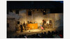 Theaterstück bei Nacht / Night-time Theatre Play Abendliche Theateraufführung im Freien mit beleuchteter Kulisse und Publikum, Hauptdarsteller:innen agieren in der Mitte der Bühne / Evening outdoor theatre performance with illuminated backdrop and audience, lead actors performing center stage