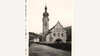 Historische Aufnahme der Kirche Flirsch mit Zwiebelturm und klassizistischer Fassade, im Vordergrund führt eine ruhige Straße entlang des Gebäudes. / Historical photograph of Flirsch Church with onion dome and neoclassical façade, a quiet road runs in front.