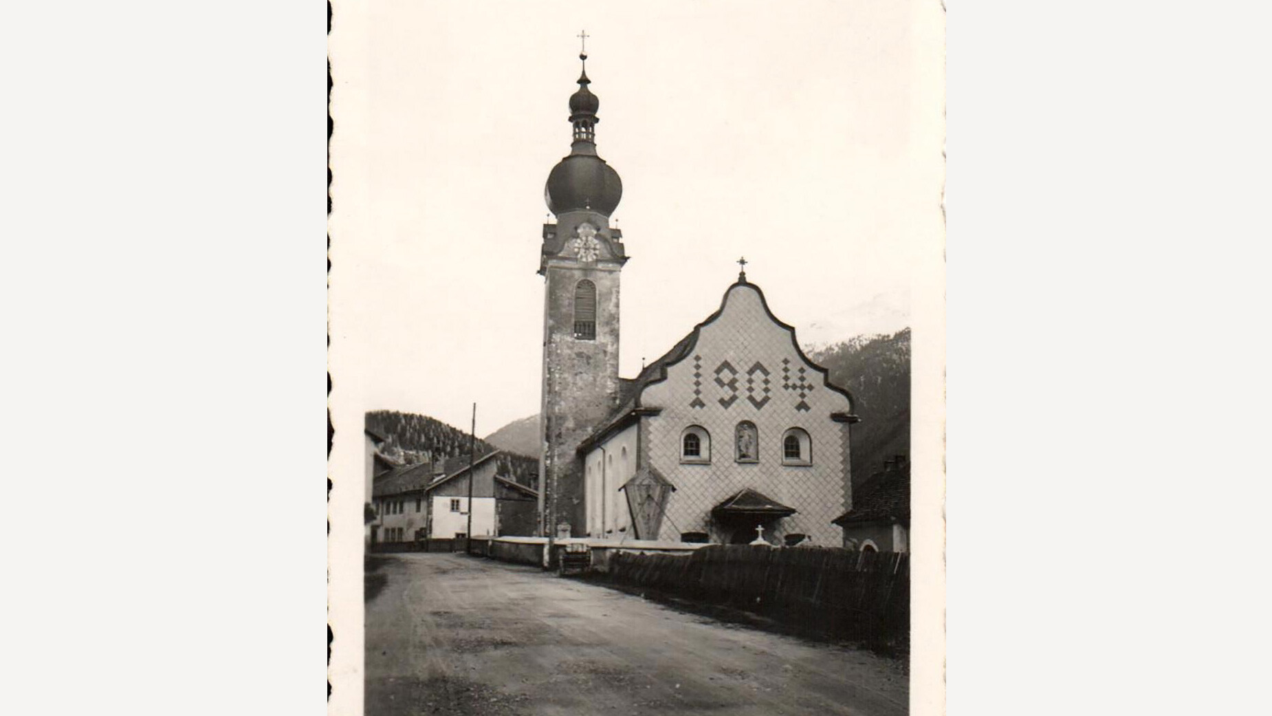 Historische Aufnahme der Kirche Flirsch mit Zwiebelturm und klassizistischer Fassade, im Vordergrund führt eine ruhige Straße entlang des Gebäudes. / Historical photograph of Flirsch Church with onion dome and neoclassical façade, a quiet road runs in front.