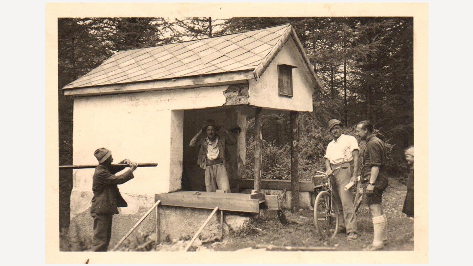 Männer bei der Restaurierung eines kleinen Kapellenbaus im Wald, Werkzeug liegt rundherum, eine Madonnafigur steht im Inneren. / Men restoring a small chapel in the forest, tools are lying around, a Madonna figure is placed inside.