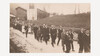 Eine Gruppe Jugendlicher in festlicher Kleidung zieht zu Fuß eine Dorfstraße entlang, im Hintergrund ein Kirchturm und ländliche Gebäude. / A group of youths in festive clothing walks along a village road, with a church tower and rural buildings in the background.