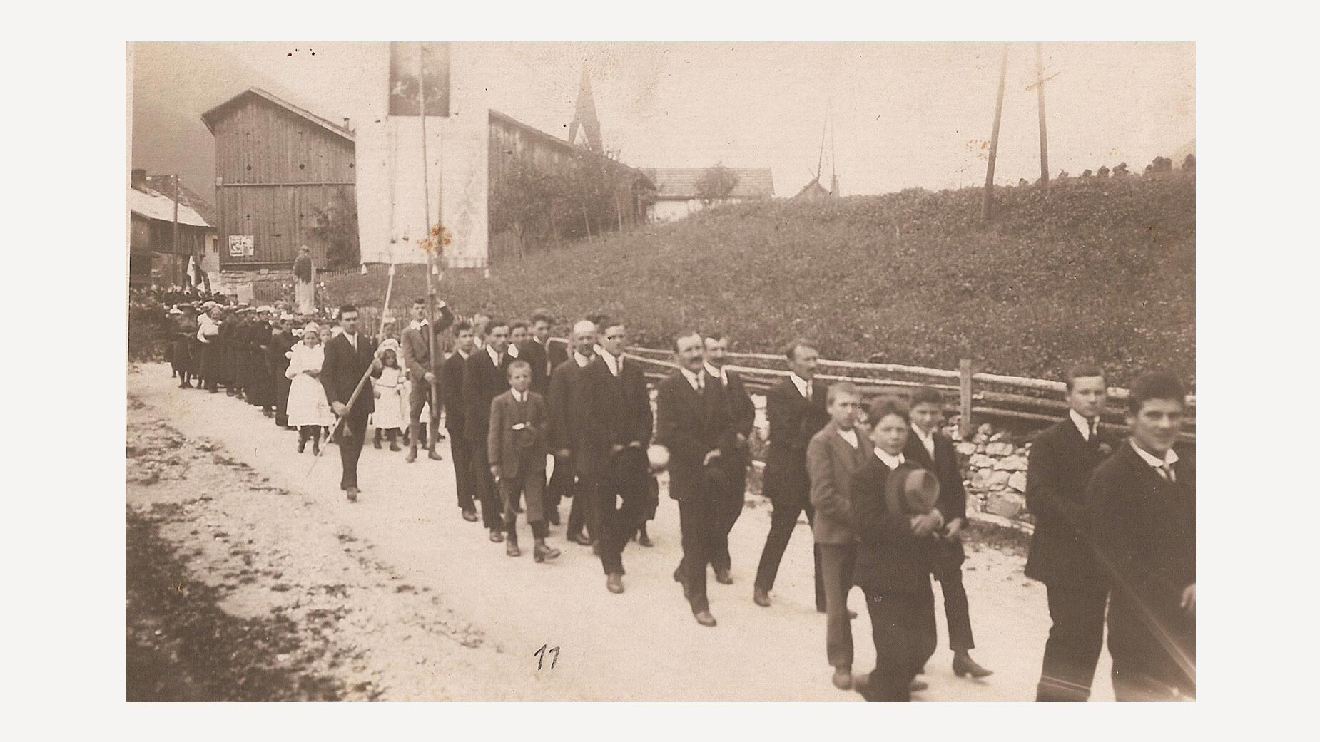Eine Gruppe Jugendlicher in festlicher Kleidung zieht zu Fuß eine Dorfstraße entlang, im Hintergrund ein Kirchturm und ländliche Gebäude. / A group of youths in festive clothing walks along a village road, with a church tower and rural buildings in the background.
