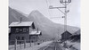 Schwarzweißaufnahme des Bahnhofs Schnann mit Gleisen, Gebäuden und markanter Bergkulisse / Black and white photo of Schnann train station with tracks, buildings and striking mountain backdrop