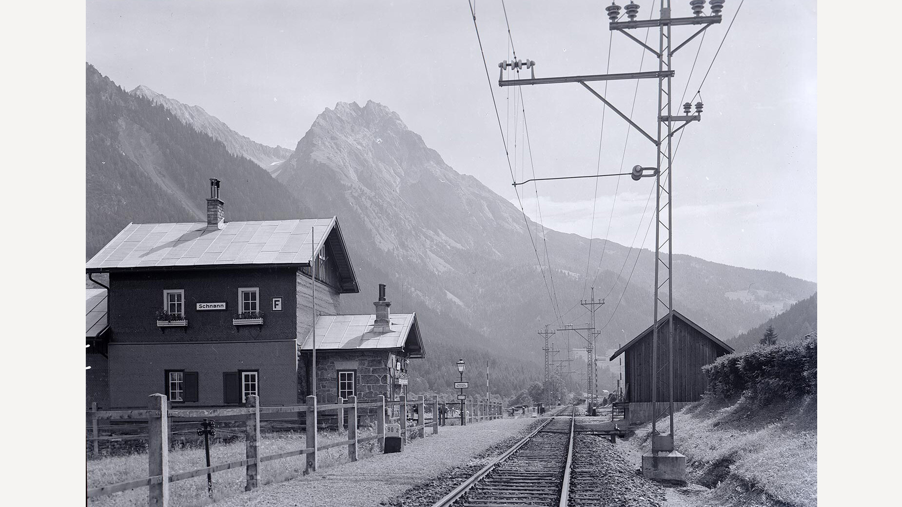 Schwarzweißaufnahme des Bahnhofs Schnann mit Gleisen, Gebäuden und markanter Bergkulisse / Black and white photo of Schnann train station with tracks, buildings and striking mountain backdrop