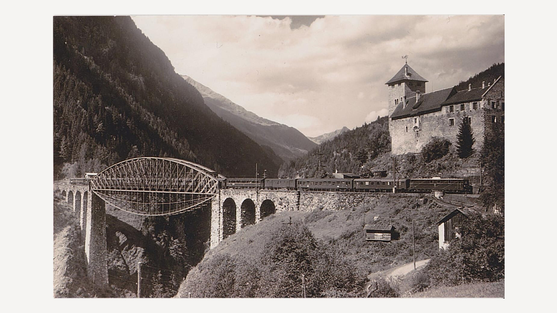 Schwarzweißaufnahme der Trisannabrücke mit vorbeifahrendem Zug und Schloss Wiesberg im Hintergrund / Black and white image of the Trisanna Bridge with a passing train and Wiesberg Castle in the background