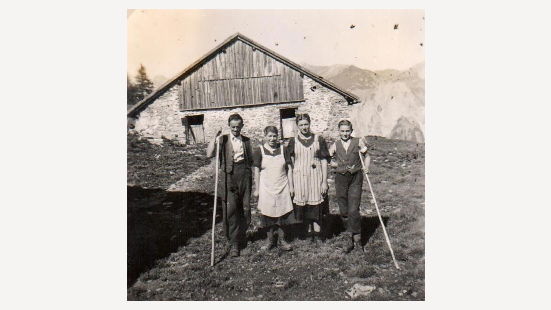 Junge Senner:innen vor Alm / Young alpine dairymen and women in front of hut Schwarzweißbild von vier jungen Menschen in traditioneller Kleidung vor einer Alm mit Werkzeugen / Black-and-white photo of four young people in traditional clothing in front of an alpine hut with tools