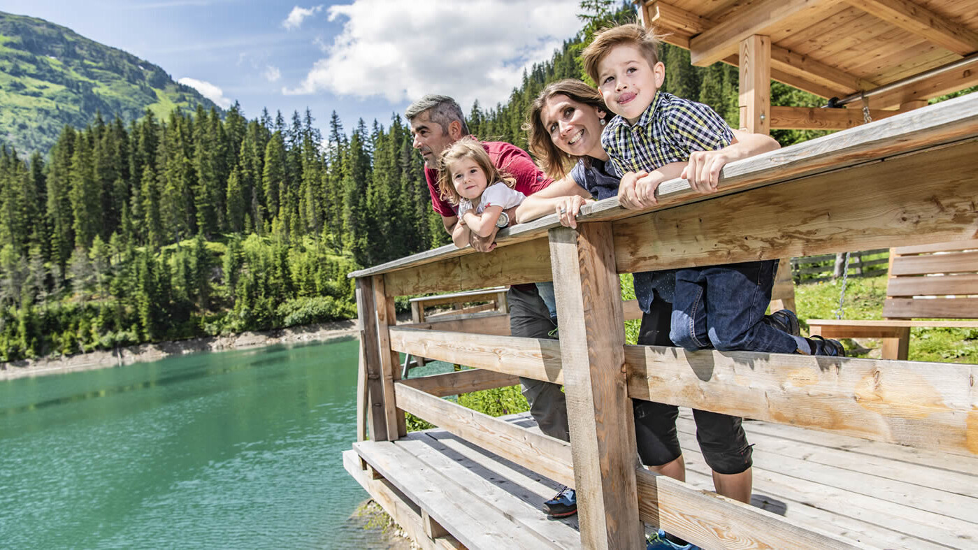 Familie lehnt sich über ein Holzgeländer an einem alpinen See mit klar grünem Wasser / Family leans over a wooden railing by an alpine lake with clear green water