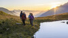 Wanderer mit Rucksäcken gehen bei Sonnenaufgang am Rand eines Bergsees entlang / Hikers with backpacks walk along the edge of a mountain lake at sunrise