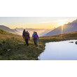 Wanderer mit Rucksäcken gehen bei Sonnenaufgang am Rand eines Bergsees entlang / Hikers with backpacks walk along the edge of a mountain lake at sunrise
