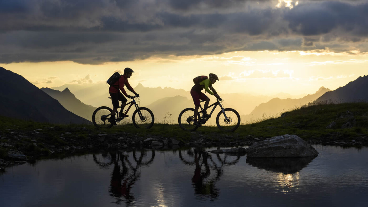 Zwei Mountainbiker fahren bei Sonnenuntergang auf einem Bergweg entlang eines stillen Sees / Two mountain bikers ride along a mountain trail by a calm lake at sunset