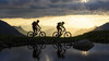 Zwei Mountainbiker fahren bei Sonnenuntergang auf einem Bergweg entlang eines stillen Sees / Two mountain bikers ride along a mountain trail by a calm lake at sunset
