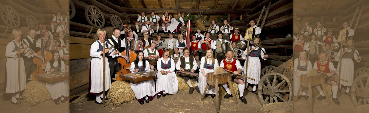 Die Volkstumsgruppe aus St. Anton am Arlberg posiert in Tracht mit traditionellen Instrumenten wie Zither, Kontrabass, Hackbrett und Blasmusik im rustikalen Stadl.