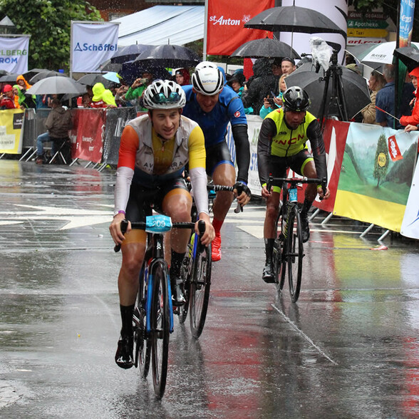 Drei Radfahrer erreichen bei Regen das Ziel, das Publikum schützt sich mit Regenschirmen.