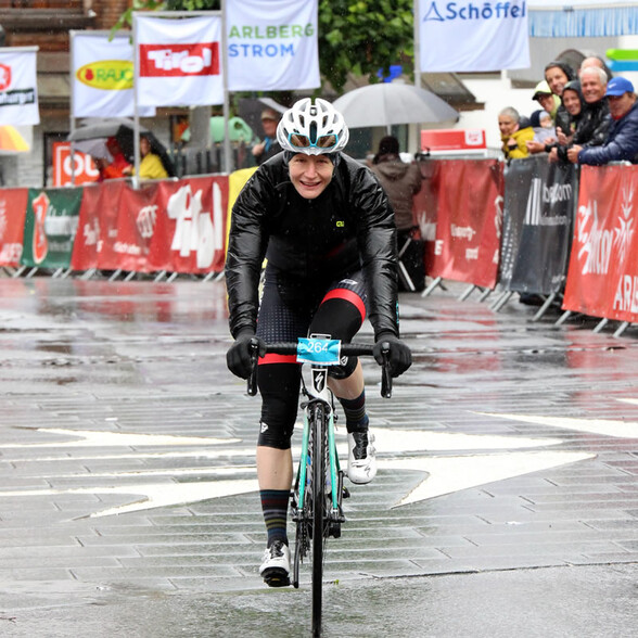 Eine Radfahrerin im schwarzen Outfit erreicht bei Regen das Ziel des Arlberg Giro, mit einem Lächeln im Gesicht.
