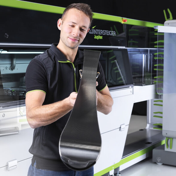 Ein Servicetechniker hält einen präparierten Ski in der Hand und begutachtet die Belagsstruktur. Im Hintergrund ist eine moderne Skiservicemaschine zu sehen.